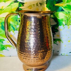 May include: A hammered copper-colored pitcher with a handle. The pitcher has a wide mouth and a rounded body. The surface has a textured, dimpled pattern. The background features green and white floral patterns.