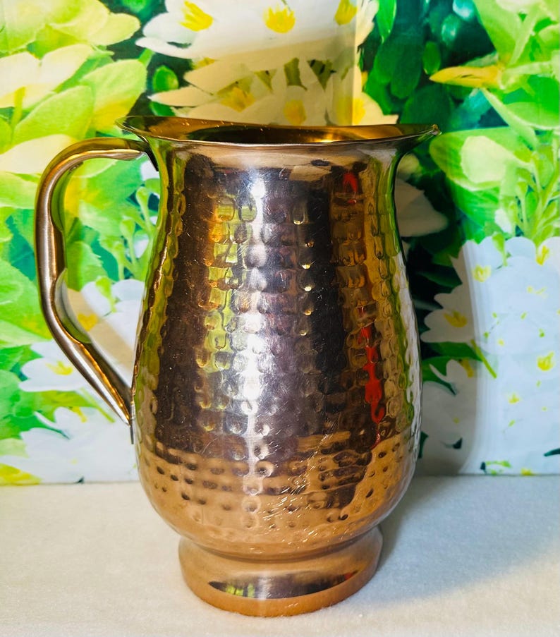 May include: A hammered copper-colored pitcher with a handle. The pitcher has a wide mouth and a rounded body. The surface has a textured, dimpled pattern. The background features green and white floral patterns.