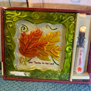 Puede incluir: Un plato decorativo con temática otoñal con un diseño de hojas en tonos naranja y amarillo, enmarcado en verde con el texto "Give Thanks to the Lord". Incluye un cuchillo pequeño con las palabras "give thanks" y una hoja.