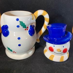 May include: A white ceramic snowman mug with a yellow and white candy cane handle. The mug is decorated with blue mittens, red berries, and green holly leaves. A separate white ceramic snowman figurine with a blue hat and yellow scarf is also included.