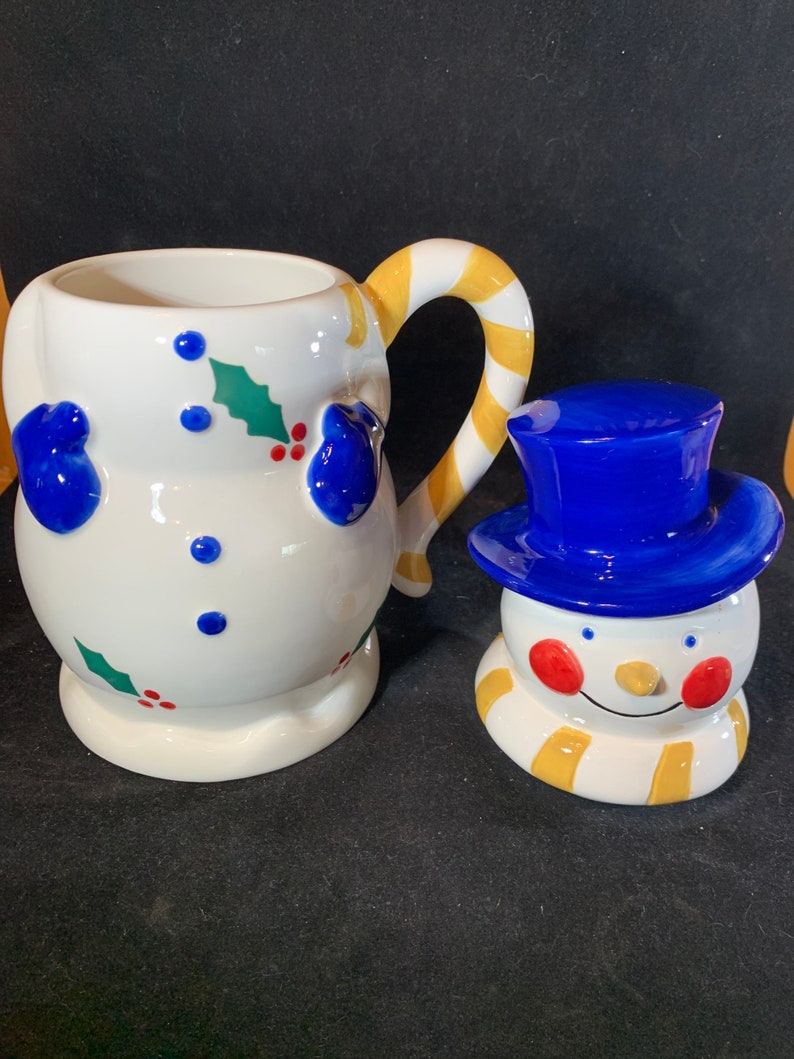 May include: A white ceramic snowman mug with a yellow and white candy cane handle. The mug is decorated with blue mittens, red berries, and green holly leaves. A separate white ceramic snowman figurine with a blue hat and yellow scarf is also included.
