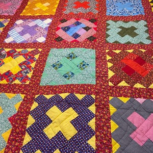 May include: A colorful patchwork quilt with a red floral border. The quilt features various square blocks in shades of yellow, blue, purple, and green. Each block has a unique pattern, creating a vibrant and cozy textile.