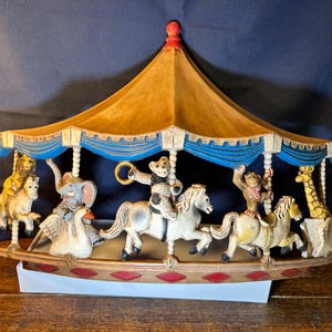 May include: A carousel figurine with a tan canopy and blue trim. The carousel features various animals, including a lion, elephant, monkey, giraffe, and swan, each riding a horse. The base is brown with red diamond accents.