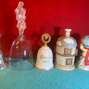 May include: Five decorative bells, each with a unique design. The bells are made of glass, porcelain, and metal. The glass bell features a clear glass body with a crystal-like figure on top. The porcelain bell has a white body with gold accents and the text "Christmas 1981". The other porcelain bell has a white body with a brown roof and windows. The metal bell has a silver body with a silver figure on top. The last porcelain bell has a white body with a blue hat and red scarf.