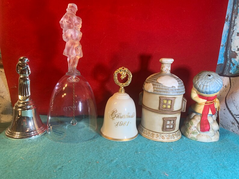 May include: Five decorative bells, each with a unique design. The bells are made of glass, porcelain, and metal. The glass bell features a clear glass body with a crystal-like figure on top. The porcelain bell has a white body with gold accents and the text "Christmas 1981". The other porcelain bell has a white body with a brown roof and windows. The metal bell has a silver body with a silver figure on top. The last porcelain bell has a white body with a blue hat and red scarf.
