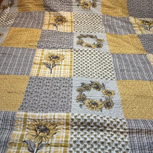 May include: A patchwork quilt featuring a repeating pattern of sunflowers, floral wreaths, and various textures. The quilt's color palette includes yellow, gray, and white, creating a cozy and inviting aesthetic. The quilt is likely made of cotton.