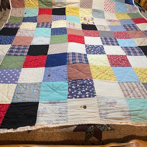 Op de afbeelding: Een patchworkquilt met een verscheidenheid aan stoffen vierkanten in rood, blauw, geel en wit. De quilt is gemaakt van verschillende patronen en texturen, waardoor een gezellig en kleurrijk ontwerp ontstaat. De quilt ligt op een bed.