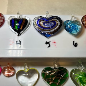 May include: A collection of heart-shaped glass pendants in various colors and designs. The pendants include swirls of blue, gold, and white, as well as green and clear glass. Each pendant has a small loop for hanging.