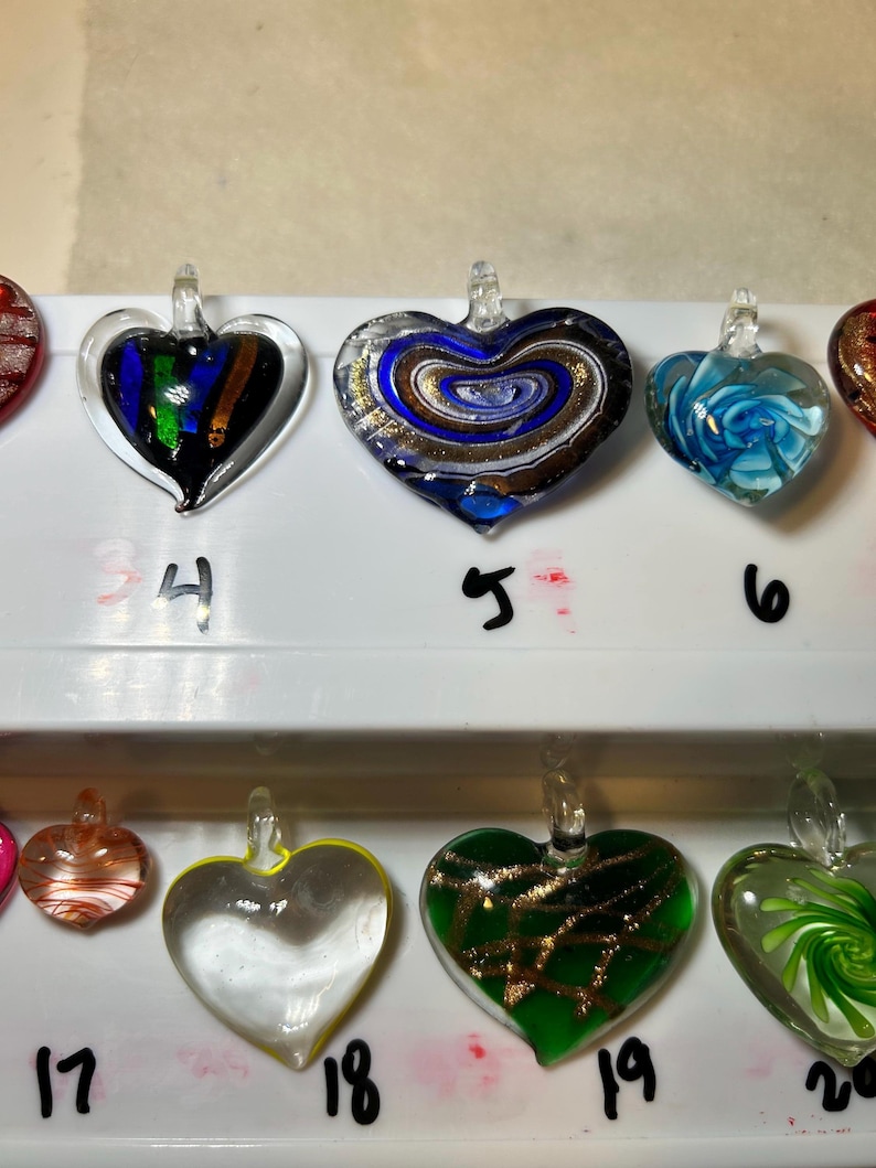 May include: A collection of heart-shaped glass pendants in various colors and designs. The pendants include swirls of blue, gold, and white, as well as green and clear glass. Each pendant has a small loop for hanging.