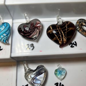 May include: A collection of heart-shaped glass pendants in various colors and designs. One pendant features a swirling pattern of pink and gray, another has gold lines on a black background, and a third has a blue butterfly.