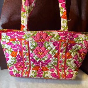 Puede incluir: Un bolso tote acolchado con un estampado floral en tonos rosa, naranja y verde sobre un fondo blanco. El bolso tiene dos asas y forma rectangular. El diseño floral se repite por toda la superficie del bolso.
