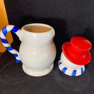 May include: A white ceramic snowman cookie jar with a red hat and blue and white striped scarf. The jar is in two pieces, the body and the head. The head is tilted to the side.