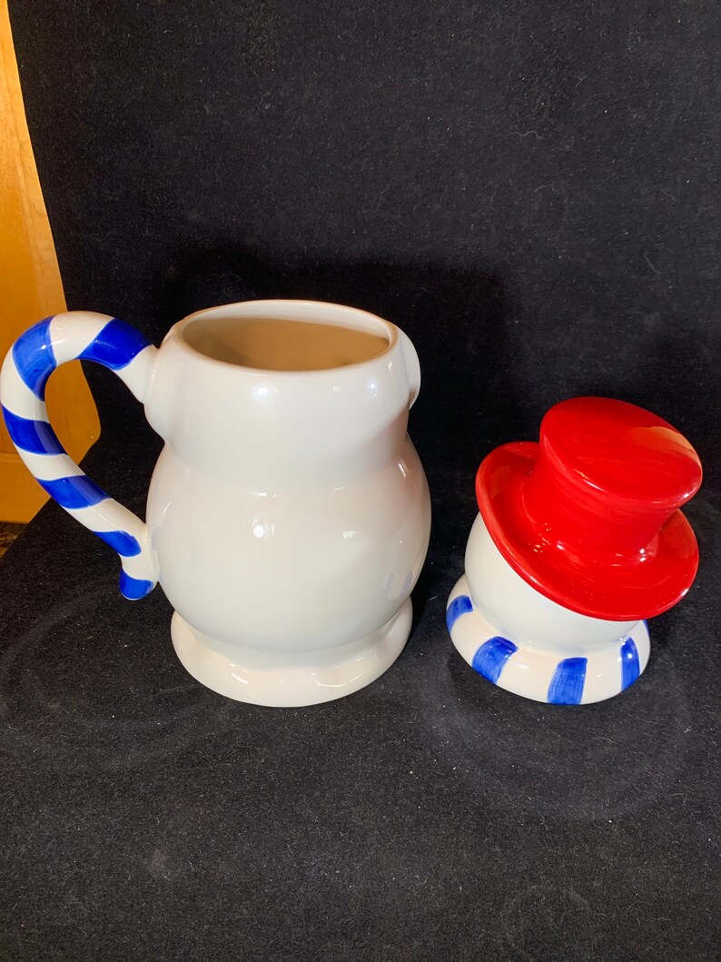 May include: A white ceramic snowman cookie jar with a red hat and blue and white striped scarf. The jar is in two pieces, the body and the head. The head is tilted to the side.