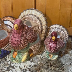 Può includere: Una collezione di statuine di tacchino in ceramica e un cestino. I tacchini hanno corpi bordeaux, motivi di piume dettagliati e sono appollaiati su basi con mais e foglie. Il cestino ha un manico ed è decorato con una testa di tacchino.