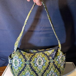 May include: A quilted shoulder bag with a navy blue background and a repeating pattern of green, blue, and white floral designs. The bag has a zipper closure and a matching patterned strap.