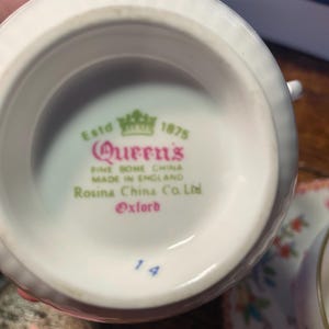 Może przedstawiać: Zbliżenie na sp&oacute;d białej filiżanki do herbaty z napisem "Queen's FINE BONE CHINA MADE IN ENGLAND Rosina China Co. Ltd Oxford" w kolorze r&oacute;żowym i zielonym. Numer "14" jest wydrukowany na niebiesko. Filiżanka jest częścią zestawu.