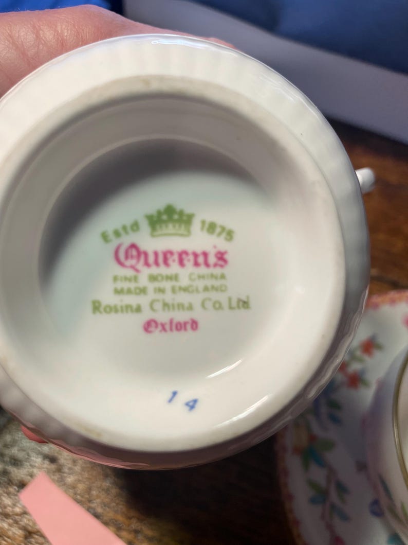Może przedstawiać: Zbliżenie na sp&oacute;d białej filiżanki do herbaty z napisem "Queen's FINE BONE CHINA MADE IN ENGLAND Rosina China Co. Ltd Oxford" w kolorze r&oacute;żowym i zielonym. Numer "14" jest wydrukowany na niebiesko. Filiżanka jest częścią zestawu.