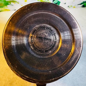 May include: Close-up of the bottom of a copper-colored mug. The center features a circular label with the text "ROBERT STANLEY SIGNATURE COLLECTION", surrounded by the words "FOOD SAFE", "HAND WASH ONLY", and "NOT MICROWAVE SAFE".
