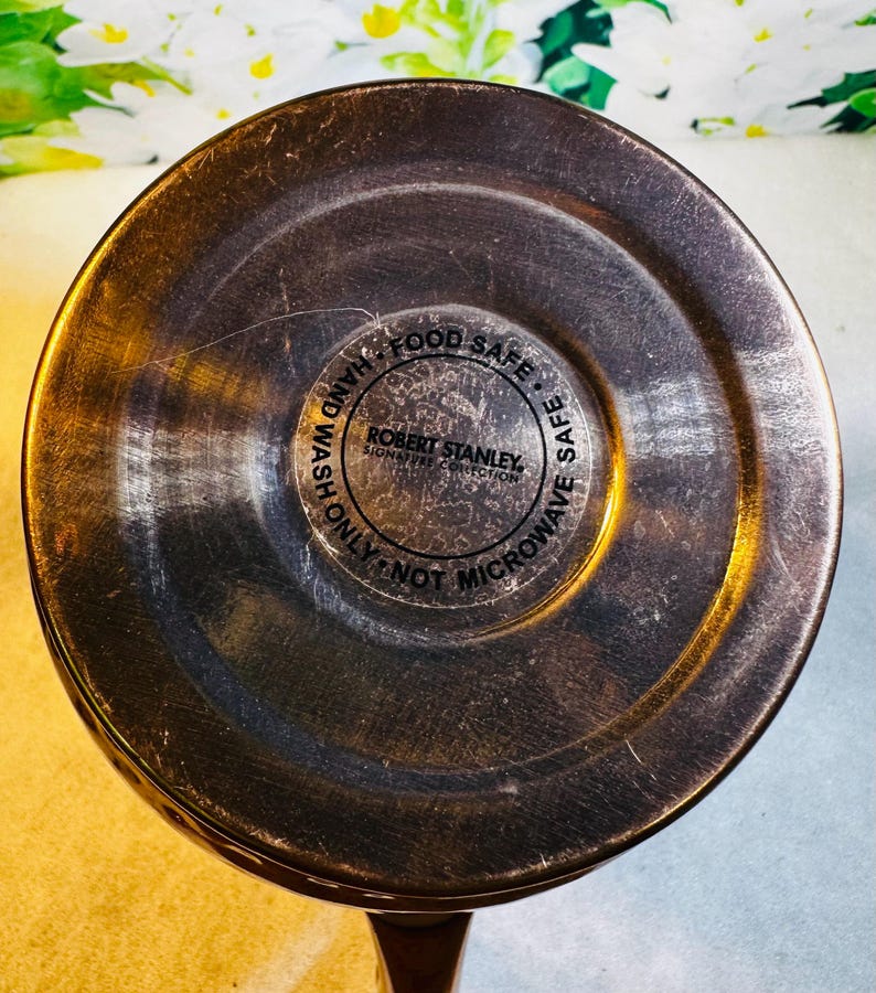 May include: Close-up of the bottom of a copper-colored mug. The center features a circular label with the text "ROBERT STANLEY SIGNATURE COLLECTION", surrounded by the words "FOOD SAFE", "HAND WASH ONLY", and "NOT MICROWAVE SAFE".