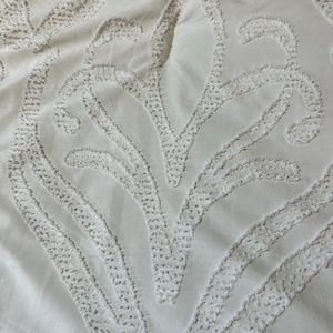 Könnte beinhalten: Nahaufnahme eines weißen Stoffes mit einem gestickten Blumenmuster. Das Design zeigt stilisierte Blattformen und geschwungene Linien, die einen strukturierten, erhabenen Effekt erzeugen. Die Stickerei ist weiß und passt zum Stoff.