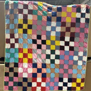 Puede incluir: Un edredón de patchwork colorido con un patrón de tablero de ajedrez. El edredón presenta cuadrados en varios colores, incluyendo rosa, azul, amarillo y blanco, con lazos rojos en cada intersección. El edredón está hecho de una variedad de telas.