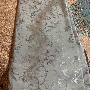 May include: A folded, pale grey fabric featuring a repeating floral pattern in silver and bronze. The fabric has a subtle sheen, and the design features stylized leaves and swirling vines. The overall aesthetic is elegant and decorative.