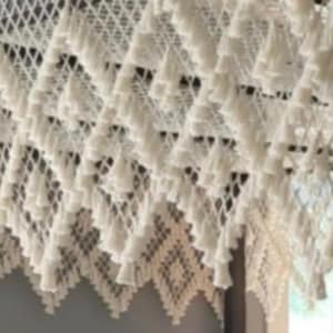May include: A white macrame wall hanging featuring a diamond-shaped grid pattern. The piece is adorned with numerous small, hanging tassels attached to the intersections of the knotted cord structure, creating a layered, textured appearance.