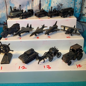 May include: A collection of 14 vintage metal pencil sharpeners, each depicting a different vehicle. The sharpeners are arranged in three rows on a white shelf. The vehicles include a boat, a train, a tank, a car, a plane, a spaceship, a motorcycle, a tractor, a carriage, and a steering wheel.
