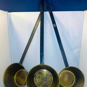 Puede incluir: Tres sartenes antiguas de latón con largos mangos negros. Las sartenes tienen forma circular con un borde ligeramente curvado. Los mangos están sujetos a las sartenes con remaches. Las sartenes están suspendidas en el aire.