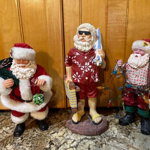May include: Three Santa Claus figurines. One Santa wears a traditional red suit and holds a sack. Another Santa is dressed in a Hawaiian shirt and sunglasses, holding a surfboard. The third Santa is in a plaid shirt and suspenders, holding Christmas lights.