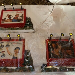 May include: Four miniature lighted billboards with red frames and white backgrounds. The billboards feature vintage advertisements for Campbell's Soup, a paint brand, and two other products. The billboards are set on a snowy base with green foliage.