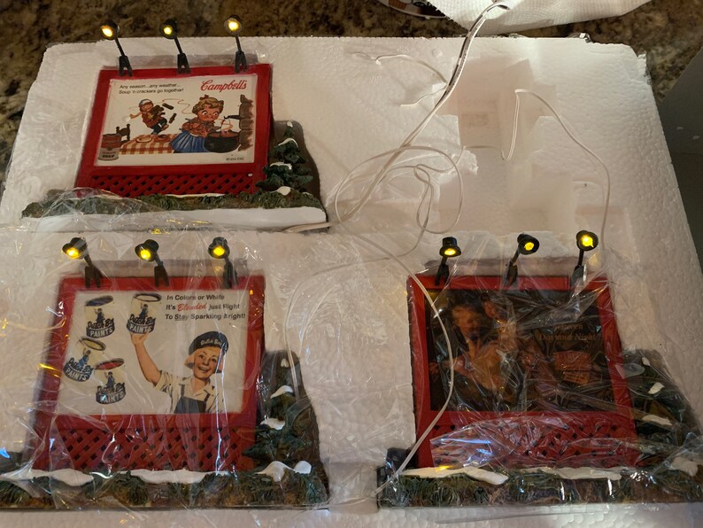 May include: Four miniature lighted billboards with red frames and white backgrounds. The billboards feature vintage advertisements for Campbell's Soup, a paint brand, and two other products. The billboards are set on a snowy base with green foliage.