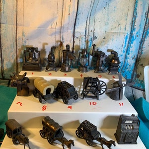 May include: A collection of 15 vintage metal pencil sharpeners in various designs, including a bell, a stove, a wagon, a windmill, and a slot machine. The sharpeners are arranged on two white shelves against a blue and white wooden background.
