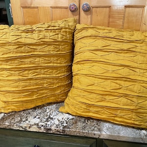 May include: Two square, mustard-yellow decorative pillows with a textured, pleated design. The pillows are made of a soft fabric and are placed on a countertop. The pillows add a pop of color and texture to any room.