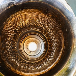 May include: An overhead view inside a metal container. The interior is a series of concentric rings, with a central light source. The metal has a tarnished, aged appearance with shades of brown and silver.