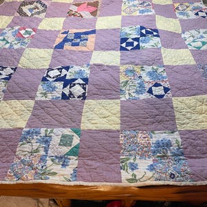 Puede incluir: Un edredón hecho a mano con un diseño de patchwork. El edredón presenta cuadrados de varios patrones y colores, incluyendo estampados florales, bloques morados lisos y crema. El edredón está extendido sobre una cama.