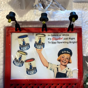 May include: A red wooden sign with a white background and a framed advertisement for Dutch Boy Paints. The sign features a young person holding a can of paint and the text "In Colors or White It's Blended just Right To Stay Sparkling Bright!"