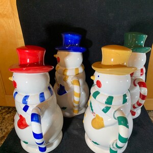 May include: Four ceramic snowman mugs with different colored hats and scarves. The mugs have a white base with colorful details and a candy cane handle.