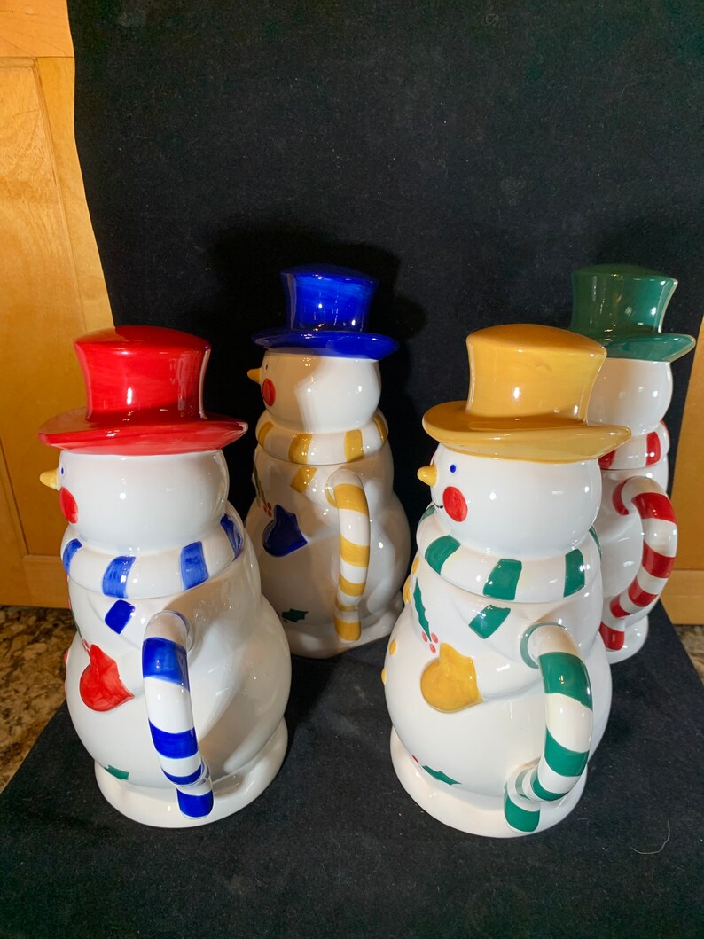 May include: Four ceramic snowman mugs with different colored hats and scarves. The mugs have a white base with colorful details and a candy cane handle.
