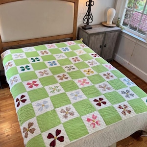 Puede incluir: Un edredón hecho a mano con un patrón de tablero de ajedrez verde y blanco. Cada cuadrado blanco presenta un diseño floral colorido en tonos de rosa, rojo, azul y marrón. El edredón está sobre una cama con cabecero de madera, creando un ambiente acogedor.
