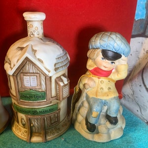 May include: A set of two ceramic Christmas ornaments. One ornament is a small house with a snow-covered roof and the words "Always Open" and "Scrooge & Marley" on the front. The other ornament is a boy wearing a blue hat and a red scarf, sitting on a rock.