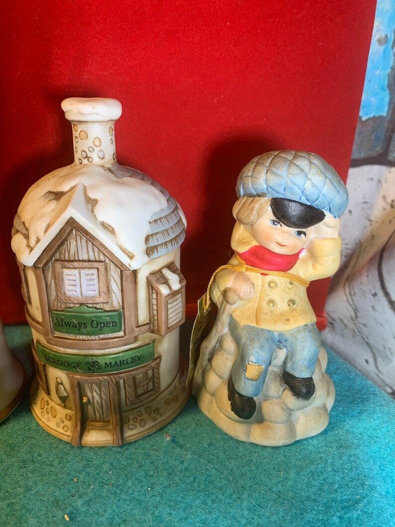 May include: A set of two ceramic Christmas ornaments. One ornament is a small house with a snow-covered roof and the words "Always Open" and "Scrooge & Marley" on the front. The other ornament is a boy wearing a blue hat and a red scarf, sitting on a rock.
