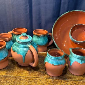 May include: A collection of handmade pottery with a rustic aesthetic. The set includes mugs, a teapot, a creamer, and a serving platter with a dip bowl. The pieces are terracotta-colored with turquoise glaze accents, creating a unique and artistic look.