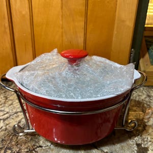 May include: A red ceramic casserole dish with a white interior and a red lid. The dish is in a silver metal stand. The dish is wrapped in bubble wrap. The casserole dish is on a speckled countertop.