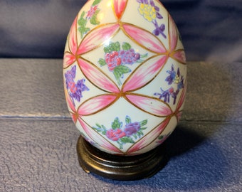 Op de afbeelding: Een wit porseleinen ei met een roze, gouden en bloemmotief. Het ei staat op een houten voet.