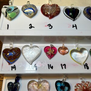 May include: A collection of heart-shaped glass pendants in various colors and designs. Some pendants feature swirls, metallic accents, or floral patterns. Each pendant has a small loop for hanging.
