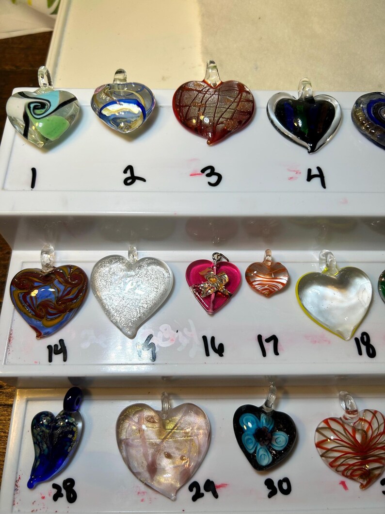 May include: A collection of heart-shaped glass pendants in various colors and designs. Some pendants feature swirls, metallic accents, or floral patterns. Each pendant has a small loop for hanging.
