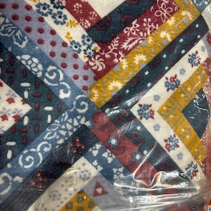 May include: A fabric quilt featuring a geometric log cabin pattern with a variety of printed designs. The textile displays a color palette of deep red, navy blue, mustard yellow, and white, with floral, polka dot, and dash motifs throughout.