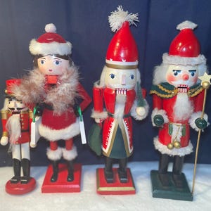 Könnte beinhalten: Vier hölzerne Weihnachtsnussknacker in festlicher Kleidung. Die Nussknacker sind in Rot, Weiß und Schwarz bemalt, mit Details wie Fellbesatz, Hüten und einem Stab mit Stern. Sie stehen auf roten und grünen Sockeln.
