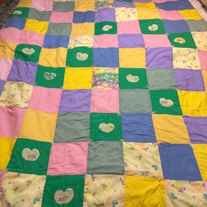 May include: A patchwork quilt in pastel shades of pink, yellow, green, blue, and purple. Some squares have heart and butterfly motifs. The quilt is displayed on a bed.